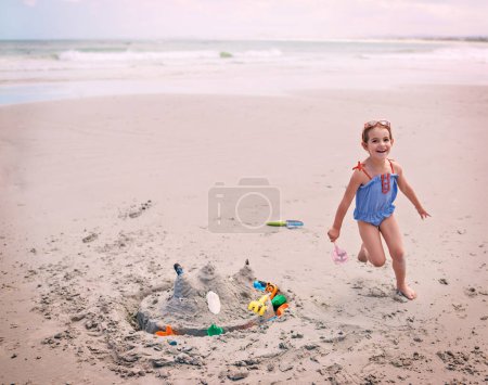Téléchargez les photos : Portrait, plage et petite fille courir avec le château de sable en plein air par la mer ou l'océan pour le plaisir insouciant. Nature, eau et énergie avec un jeune enfant heureux jouant sur le sable de la côte pour des vacances ou des vacances. - en image libre de droit