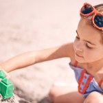 Girl, toys and building sandcastle on the beach with shape or block for fun summer, holiday or weekend in nature. Female person, child or kid playing and enjoying sand construction by the ocean coast.
