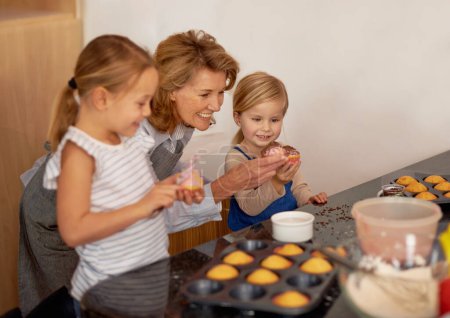 Photo for Grandmother, child and cupcake helping in kitchen or icing decorations or sprinkles or sweet treat, bonding or teaching. Female person, siblings and baking ingredients or learning, dessert or recipe. - Royalty Free Image