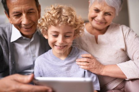 Téléchargez les photos : Famille heureuse, grands-parents et enfant avec tablette pour le divertissement, les médias sociaux ou la recherche sur le canapé à la maison. Grand-mère, grand-père et petit garçon sourient sur la technologie pour la recherche en ligne ou film à la maison. - en image libre de droit