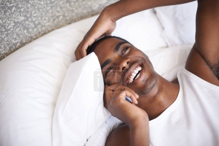 Foto de Feliz, hombre negro y acostado en la cama con teléfono para la comunicación, conversación o redes en casa. Cara de hombre africano con sonrisa en el teléfono inteligente móvil para la discusión o relajarse en el dormitorio. - Imagen libre de derechos