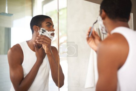 Téléchargez les photos : Miroir, rasage ou toilettage et homme noir dans la salle de bain de la maison avec rasoir pour l'épilation du matin. Visage, soins de la peau et réflexion des jeunes avec de la mousse pour raser la barbe pour la routine d'hygiène. - en image libre de droit