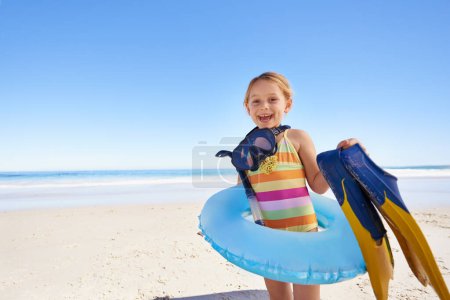 Foto de Playa, niño y vacaciones para nadar en verano con retrato en un ambiente tranquilo. Chica, sonrisa y persona emocionada en la arena con traje, inflable y gafas de sol para la seguridad y la precaución. - Imagen libre de derechos