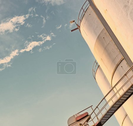 Photo for Closeup, silo and tank at plant for agriculture, bulk storage and product safety with sky, clouds and outdoor. Container, agricultural structure and oil refinery tower with low angle for distillery. - Royalty Free Image