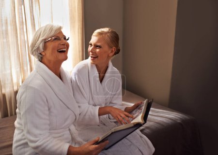 Photo for Grandmother, woman and laughing at photo album, home and happy for bonding and looking at memories. Senior lady, daughter and moment for self care, robe and bedroom for comfort and hospitality. - Royalty Free Image