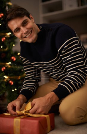 Téléchargez les photos : Portrait, arbre de Noël et l'homme emballage cadeau à la maison pour la préparation de l'événement de vacances de la tradition. Cadeau, cadeau et sourire avec jeune heureux dans l'appartement pour la célébration de décembre ou des vacances. - en image libre de droit
