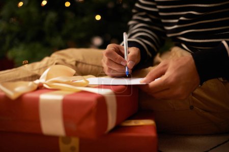 Téléchargez les photos : Stylo, mains et homme avec carte et cadeau pour l'événement de Noël ou fête à la maison pour la famille. Célébration, papier et gros plan de la lettre d'écriture de la personne masculine avec des boîtes-cadeaux pour Noël vacances festives - en image libre de droit