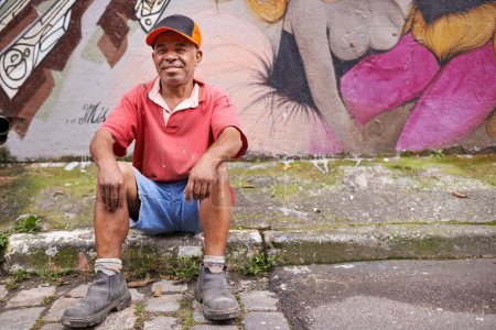 Téléchargez les photos : Sans-abri, homme et pauvreté dans la rue, homme mûr et art du graffiti sur le mur en zone urbaine. Sourire, assis et trottoir dans le quartier, le tiers monde ou design créatif en communauté pour se détendre. - en image libre de droit