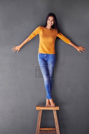 Photo for Woman, clothes and stand on chair in portrait, smile and fashion on wall background. Female person, cool and happy lady on stool, confident and apparel in living room at home in house for trends. - Royalty Free Image