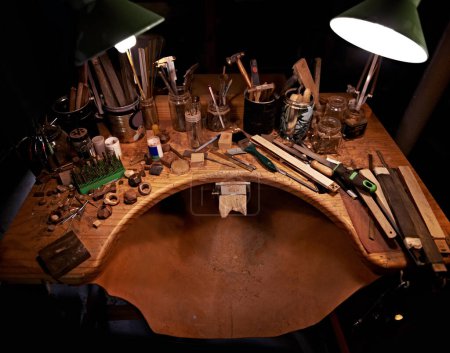 Foto de Trabajos de madera, herramientas y mesa en taller para artista en escultura para iniciar proyecto por la noche. Espacio de trabajo oscuro, estudio y creativo para carpintero con equipo en el escritorio para el proceso y la producción. - Imagen libre de derechos