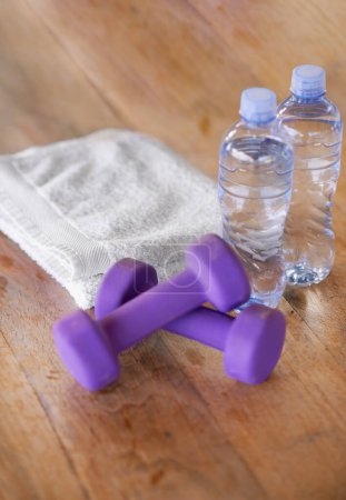 Photo for Closeup, water bottles and towel with dumbbells, ground and exercise with equipment and nutrition. Weights, h2o and cloth with wooden floor or fitness with workout or training with wellness or health. - Royalty Free Image