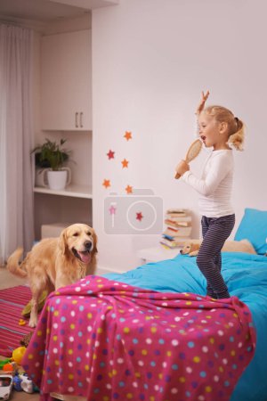 Foto de Chica, niño y cachorro con jugar en la cama para karaoke, vinculación y canto en el dormitorio del hogar con cepillo. Niño, golden retriever y perro para acompañar, bailar y relajarse con el cuidado de mascotas en el apartamento. - Imagen libre de derechos