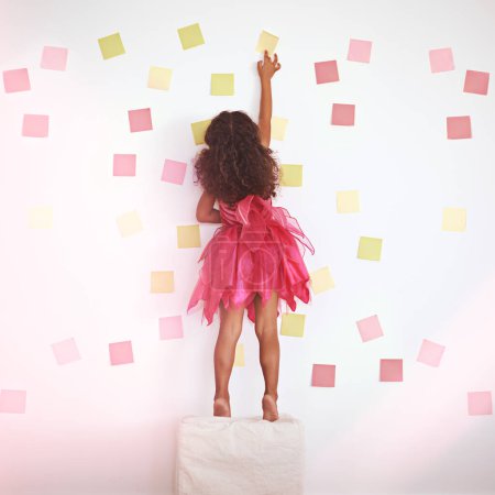 Photo for Child, girl and against wall to play with sticky notes, colors and creativity with standing on ottoman. Kid, female person and home with childhood activity for development, growth and cute in dress. - Royalty Free Image