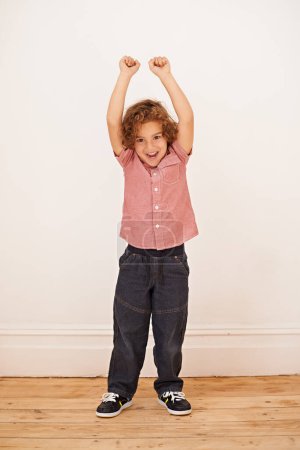Photo for Excited boy, child and portrait with fist in air by wall in family home for game, winning or success. Kid, happy and playful with smile for victory, cheers and celebration for fun in living room. - Royalty Free Image