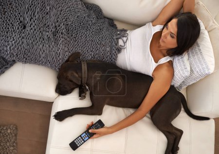 Remote control, woman on a couch and relaxing with a dog and peaceful with weekend break and home. Person, pet owner and girl watching television in a living room with morning show and best friend.