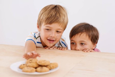 Photo for Children, boys and smile for cookies in home with stealing, peeking and childhood fun in at dining room table. Siblings, kids and happy for biscuits, snack and baked goods in kitchen of apartment. - Royalty Free Image