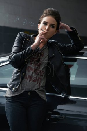 Foto de Mujer, esperando y fumando cigarrillo en el coche en el estacionamiento para recoger o transporte. Persona femenina, fumadora o conductora de moda con chaqueta de cuero e inhalación de tabaco por vehículo estacionario. - Imagen libre de derechos