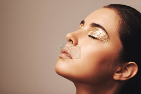 Photo for Woman, face and gold glitter on eyes for beauty, cosmetology and glamour with cosmetics on beige background. Eyeshadow, makeup and fashion model in studio, art or creativity with shimmer and glow. - Royalty Free Image