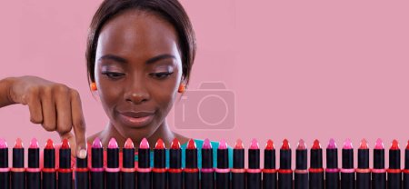 Téléchargez les photos : Rouge à lèvres, maquillage et choix de couleur avec femme noire en studio isolé sur fond rose pour les cosmétiques. Visage, vente au détail et achats de produits de beauté avec un jeune client sur l'espace de décision. - en image libre de droit