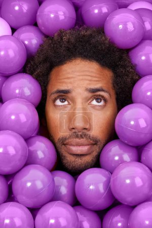 Photo for Ball pit, thinking and face of black man with plastic toys for wondering, question and thoughtful on background. Confused, facial expression and person with purple balls, decoration and objects. - Royalty Free Image