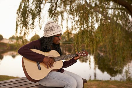 Photo for Young woman, playing and guitar on bench by lake for music, passion and talent on string instrument. African lady, musician and composer of song in nature and jazz artist with practice for concert. - Royalty Free Image