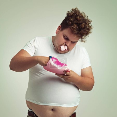 Photo for Food, hunger and marshmallow with plus size man in studio on gray background for unhealthy eating. Belly, hungry for sweets and candy with young person with snack bag or packet for greed or gluttony. - Royalty Free Image