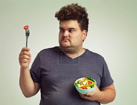Photo for Plus sized, man and unhappy with food for healthy diet, fork or bowl and salad for wellness on studio background. Male person, detox and lose weight for health, eating disorder and nutrition. - Royalty Free Image