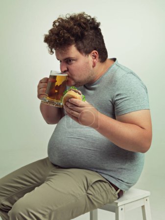 Photo for Man, drink and beer with burger in studio for unhealthy diet, fast food and nutrition on chair. Plus size, hungry and male person with alcohol on stool for dinner, meal and beverage in glass. - Royalty Free Image