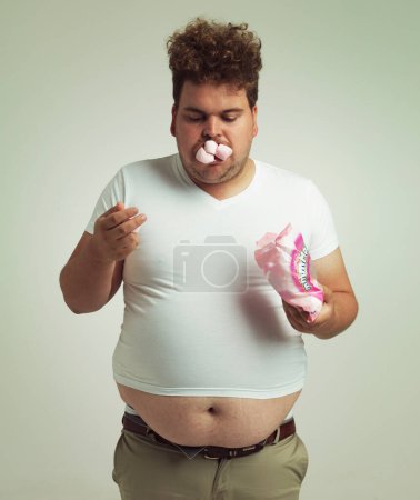 Photo for Obesity, weight gain and humor for man in studio with marshmallow for unhealthy eating, sugar and candy. Overweight person with sweets in mouth and fat stomach for greed, comedy and funny indoor. - Royalty Free Image
