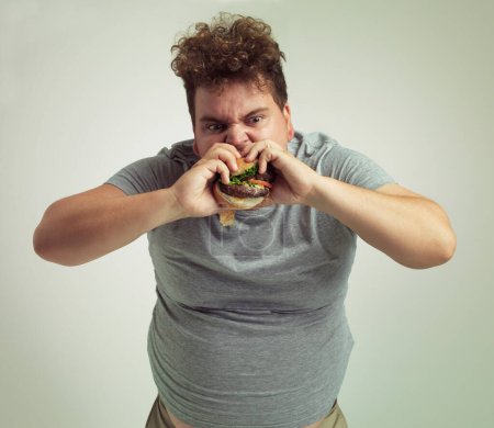 Photo for Takeaway, weight gain and burger for man in studio with fastfood for unhealthy eating, plus size and greed. Young person with meal in mouth and fat stomach for hunger, meat and sauce for bite indoor. - Royalty Free Image
