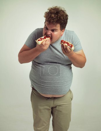 Photo for Plus size man, fast food and eating hotdogs for lunch in studio background for snack, hunger and craving. Male person, hungry and takeaway meal with ketchup or starving and enjoy while standing. - Royalty Free Image