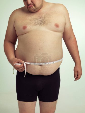 Téléchargez les photos : Homme, plus la taille et déçu sur le ventre avec mesure en studio sur fond blanc avec ruban adhésif pour la perte de poids et le progrès. Tristesse, forme physique et corps avec auto-soin pour surveiller la santé et la forme - en image libre de droit