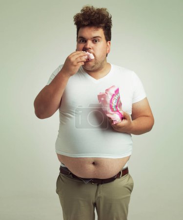 Photo for Plus size, candy and portrait for man in studio with marshmallow for unhealthy eating, sugar and confection. Young person with sweets in mouth and round stomach for greed, comedy and humor indoor. - Royalty Free Image