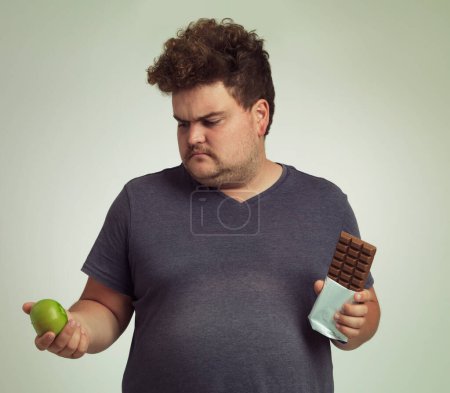 Photo for Man, apple and chocolate in studio with choice for diet, healthy food and nutrition for wellness. Male person, plus size and thinking with decision for fruit, sweet dessert and snack for lifestyle. - Royalty Free Image