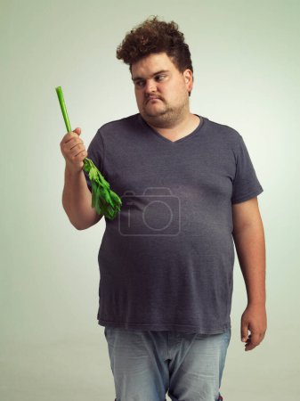 Photo for Plus size, diet and unhappy man with vegetable for health, nutrition and wellness in studio in white background. Weight loss, celery and frustrated male person for healthy food, wellbeing and detox. - Royalty Free Image
