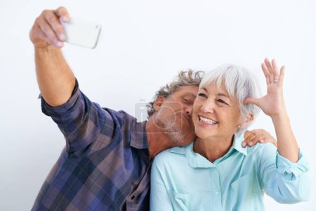 Photo for Old couple, kiss in selfie and happy together for social media post, memory and love with fun on white background. Affection, trust or loyalty with people in marriage, smile in picture and mobile app. - Royalty Free Image