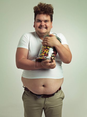 Photo for Candy, crazy and portrait of plus size man in studio for luxury snacks, sweets and dessert in container. Comic, funny facial expression and isolated person with glass for unhealthy diet on background. - Royalty Free Image