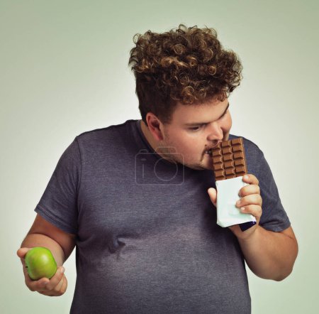 Photo for Man, apple and chocolate in studio with choice of junk food, sweet or diet for wellness. Plus size, male person eating and decision, fruit or candy for nutrition, lose weight and healthy lifestyle. - Royalty Free Image