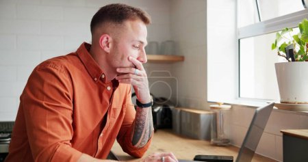 Photo for Man, thinking and typing on laptop at home, planning and reflection by kitchen window in apartment. Male person, freelancer and contemplating ideas or online research, website and remote work or job. - Royalty Free Image