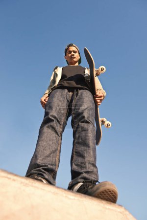 Skatepark, serious and portrait of man with skateboard practicing for competition with skills. Fitness, hobby and bottom view of young male skater on ramp for outdoor training with blue sky