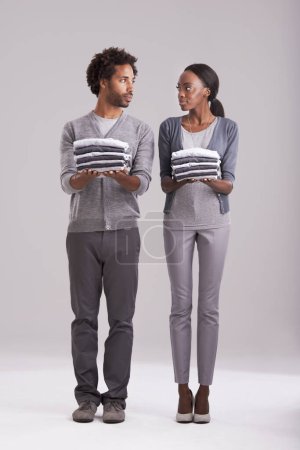 Photo for Couple, laundry and teamwork in studio for helping, support and domestic chores on a white background. Serious African man and young woman with clothes in hands for fashion, neat and gender equality. - Royalty Free Image