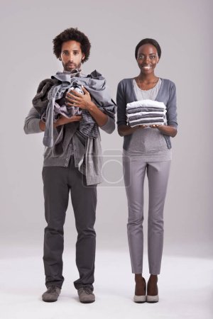 Photo for Portrait, studio and people with pile of clothes in hand for laundry day, wash or cleaning together. Woman, smile and washing folded with messy, man and annoyed for dirty wardrobe on gray background. - Royalty Free Image