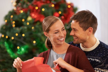 Téléchargez les photos : Amour, cadeau de Noël et couple dans le salon à la maison pour la célébration festive ensemble. Sourire, heureux et jeune homme et femme avec boîte cadeau de vacances pour l'événement de Noël ou fête à la maison moderne - en image libre de droit
