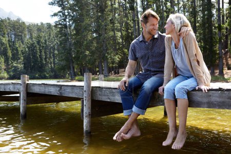 Téléchargez les photos : Couple, lac et assis sur la jetée, en plein air et heureux pour étreindre en vacances, se détendre ou la nature en été. Homme, femme et embrasser avec sourire, soins ou lien avec la connexion pour l'amour en vacances dans le Colorado. - en image libre de droit