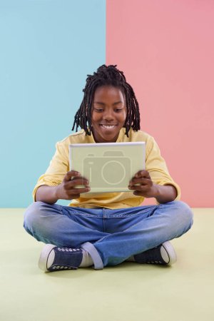 Téléchargez les photos : Adolescent, lecture et tablette en studio pour l'éducation en ligne, e-learning et jeux créatifs sur le sol. Enfant, étudiant ou garçon africain avec technologie numérique en couleur pastel ou fond bleu et rose. - en image libre de droit