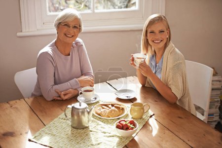 Téléchargez les photos : Portrait, femme heureuse et mère âgée buvant du thé au petit déjeuner, collant et sourire à la maison. Visage, maman aînée et adulte avec tasse à café à table, nourriture et biscuits familiaux à la maison ensemble. - en image libre de droit