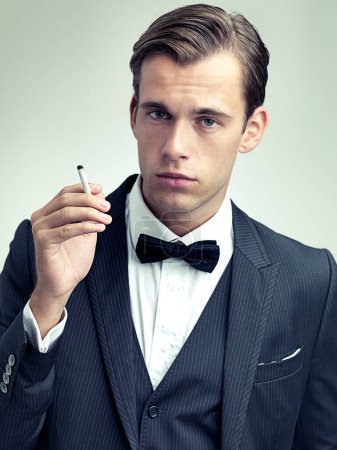 Photo for Portrait, elegant or young gentleman smoking a cigarette in studio, confidence or vintage fashion by white background. Face, serious and man in retro suit or tuxedo, bow tie and class at formal event. - Royalty Free Image