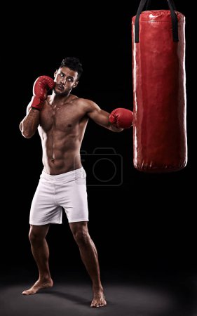 Téléchargez les photos : Studio, athlète et homme avec sac de boxe et gants pour l'entraînement avec force et muscle. Adulte, boxeur et homme dans un fond sombre, sain et puissant avec le corps pour le sport et sérieux pour le travail. - en image libre de droit