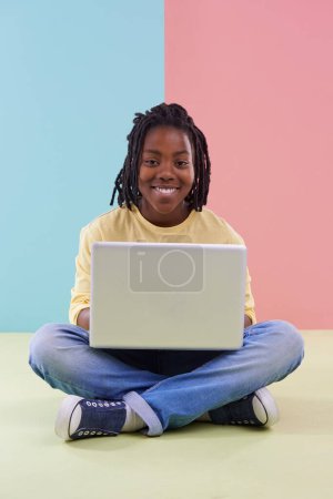 Photo for Teenager, portrait and laptop in studio for online education, e learning and creativity on the floor. Kid, student or African boy with computer for school in pastel color or blue and pink background. - Royalty Free Image