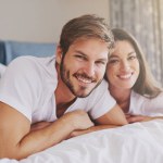 Happy, hotel and portrait of couple on bed for relaxing, bonding and resting together on vacation. Smile, romantic and young man and woman laying in bedroom at modern apartment or home on weekend